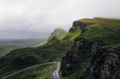 Winding single-track road through misty green Scottish highland cliffs under an overcast sky