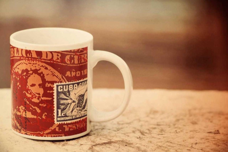 White ceramic coffee mug printed with a Cuban postage stamp design, resting on a marble surface
